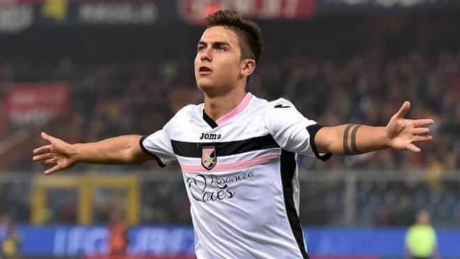 Paulo Dybala pictured in Palermo's mainly white away kit, with a pink and a black horizantal stripe across the chest. He is running with his arms raised. 